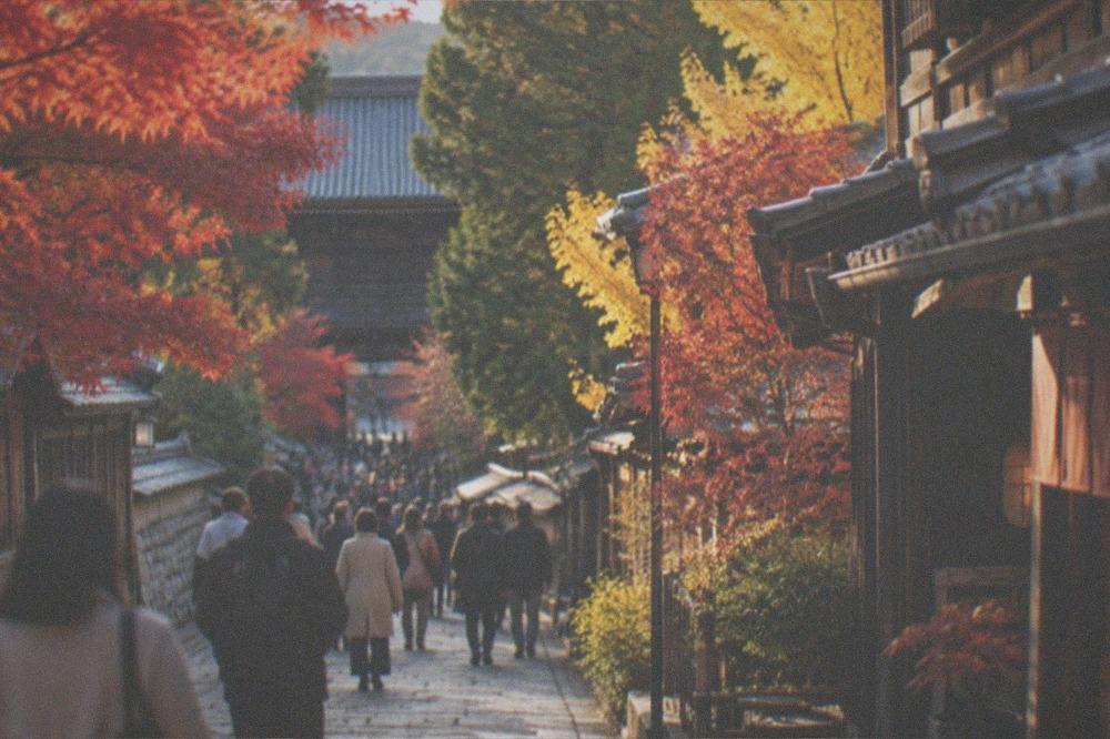 A crowded stone lane in Kyoto lined with maple trees and traditional wooden shopfronts