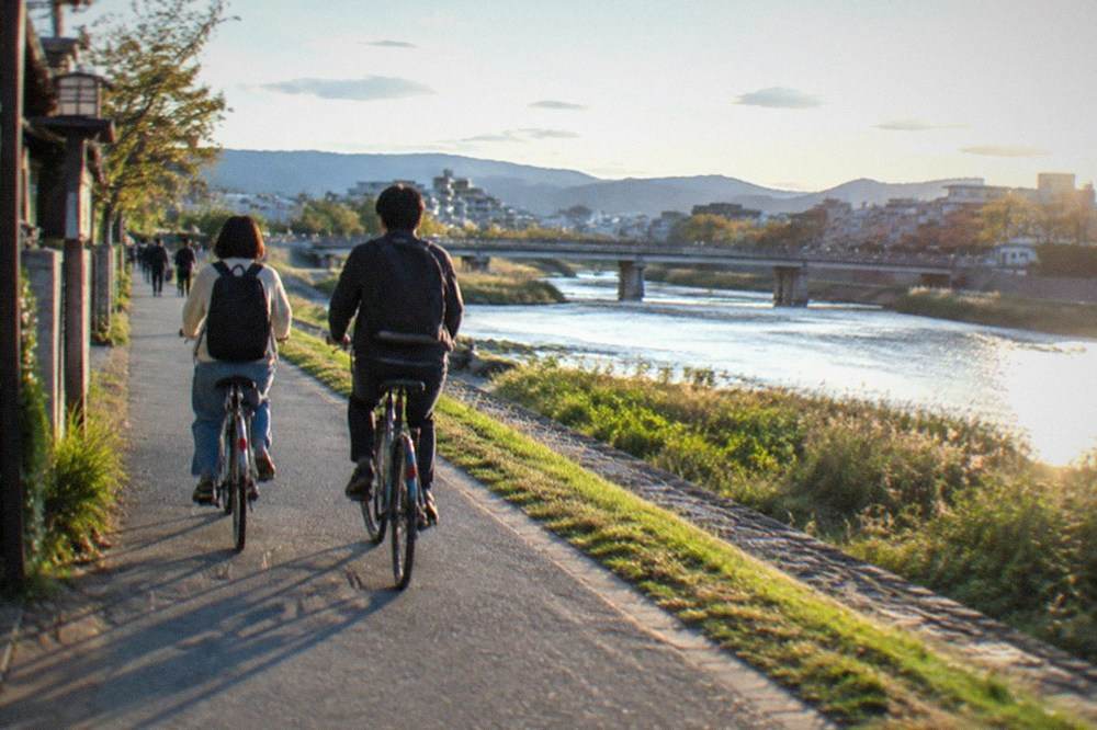 Two cyclists riding side by side on a paved path beside the Kamo River in Kyoto