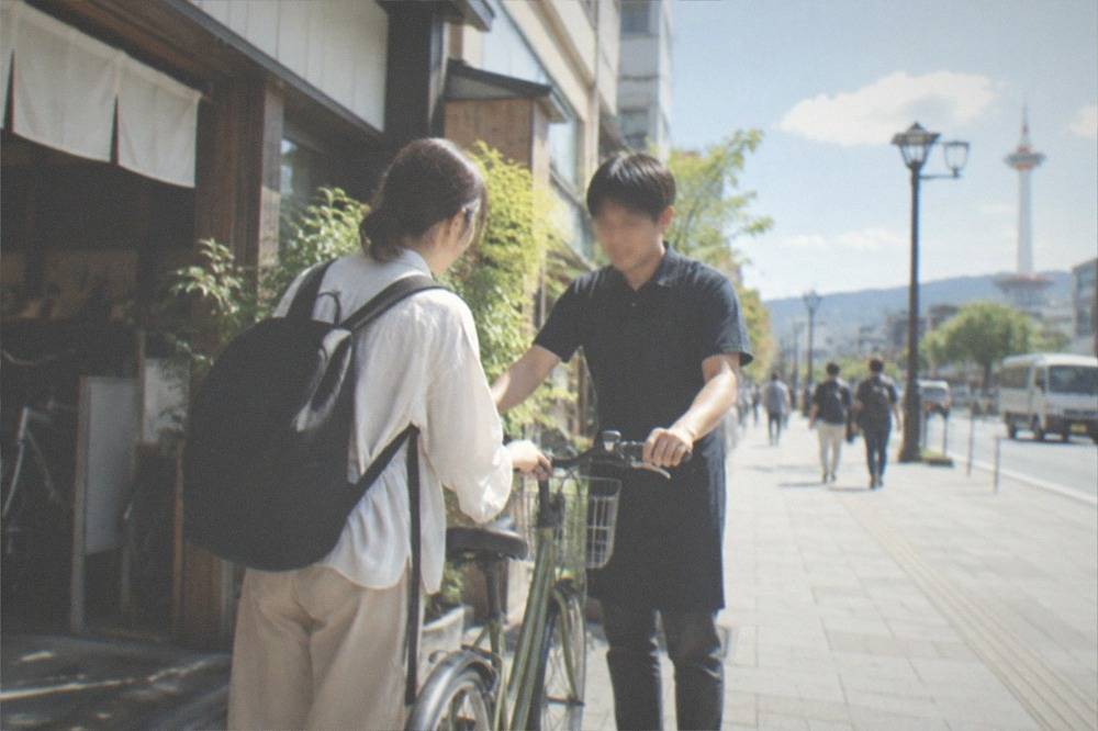 A rental shop employee handing a bicycle to a customer outside a small Kyoto storefront