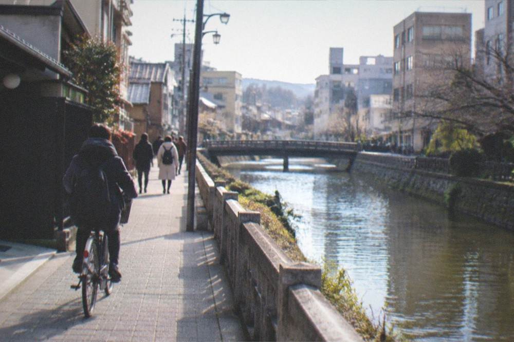 Cyclists and pedestrians on a narrow street beside a Kyoto canal