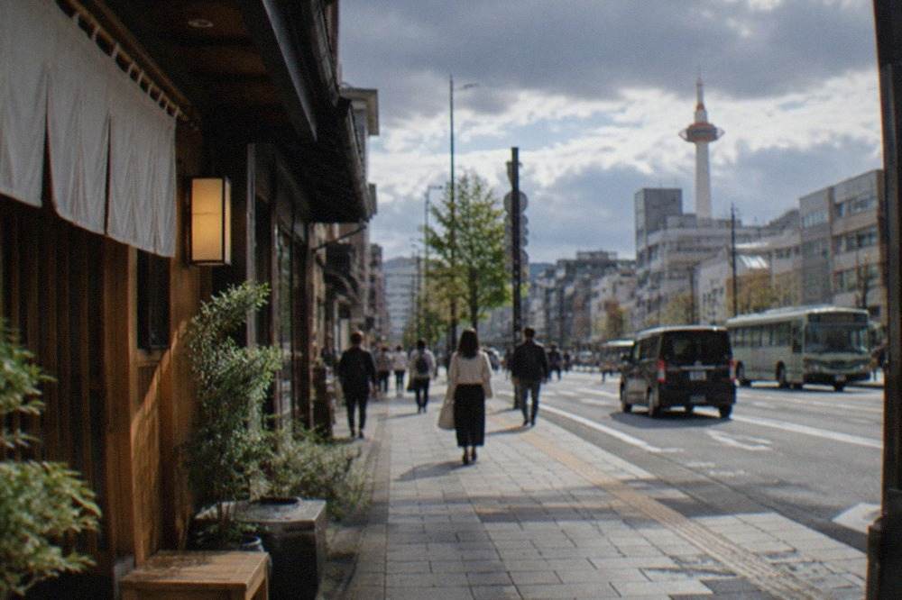 A green Kyoto City Bus stopped at a busy street stop with passengers waiting to board