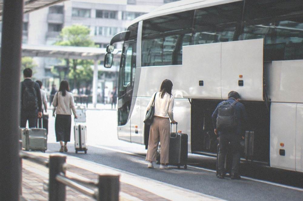 A white airport limousine bus parked at a terminal curb with passengers loading luggage