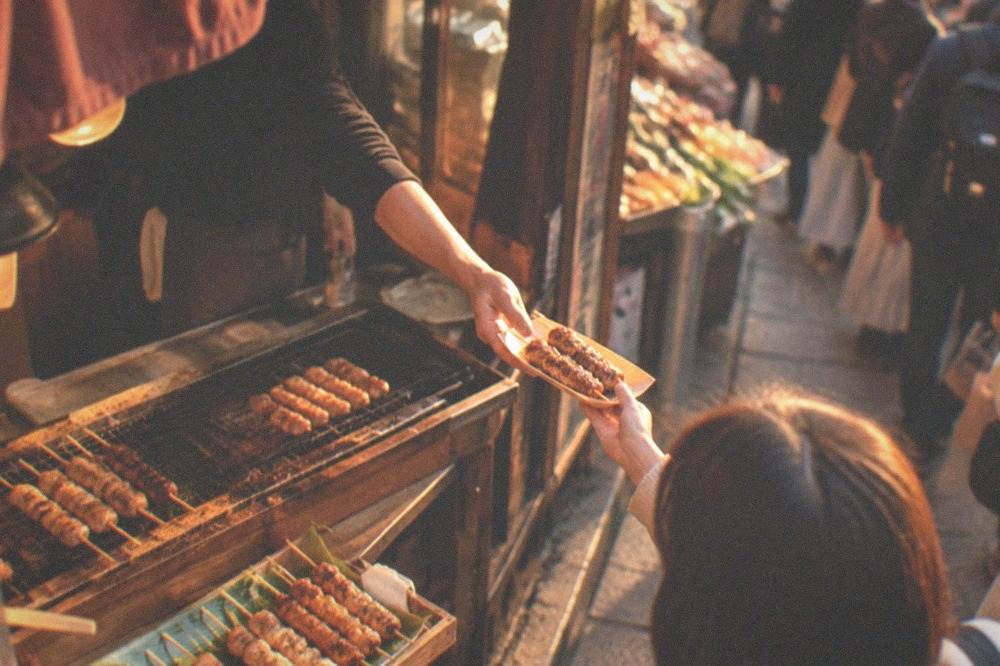 A cook handing grilled food across a busy Nishiki Market stall counter