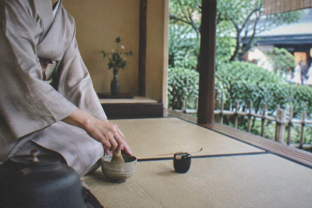 A host whisking matcha in a ceramic bowl during a tea ceremony in a tatami room