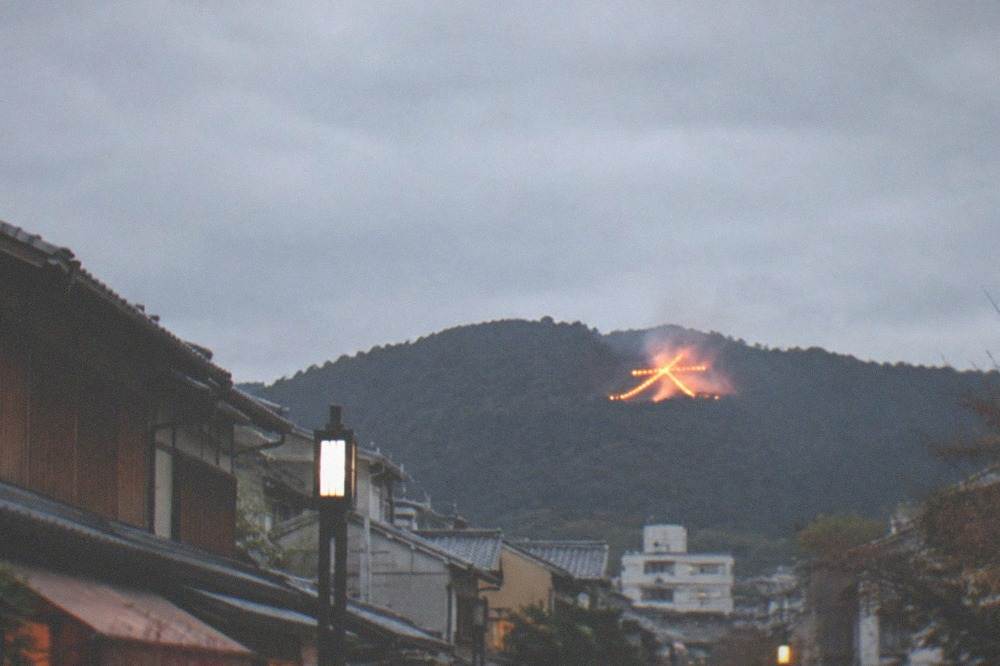 A giant bonfire character glowing on a dark mountain above Kyoto rooftops