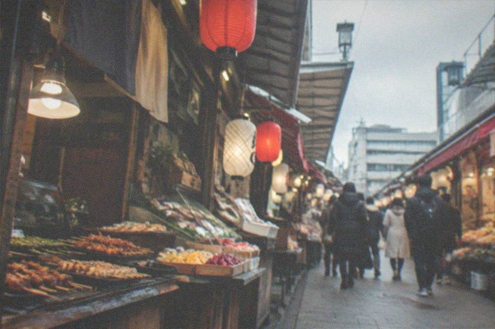 A narrow covered market lane lined with food stalls selling skewers, pickles and fish under hanging lanterns