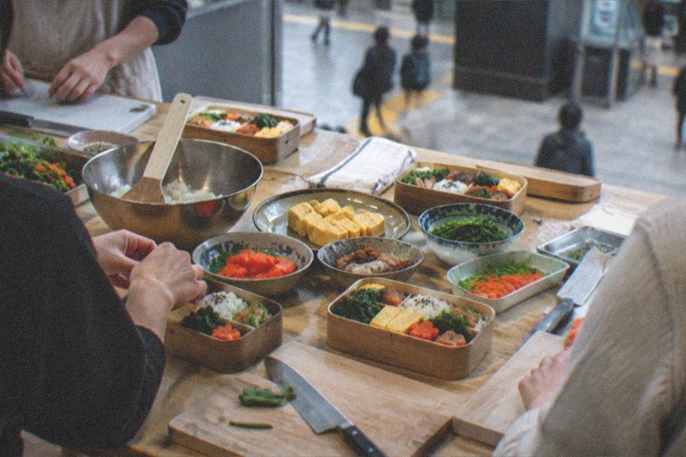 A cooking class table with ingredients, knives and half-prepared bento boxes laid out for students