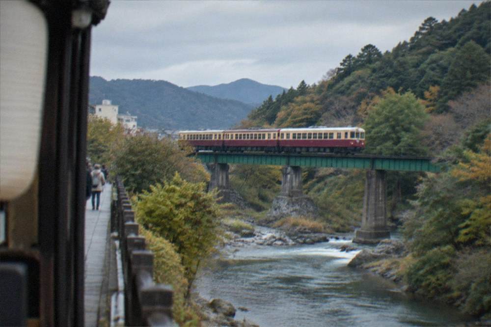 A small vintage sightseeing train crossing a wooded river valley north of Kyoto