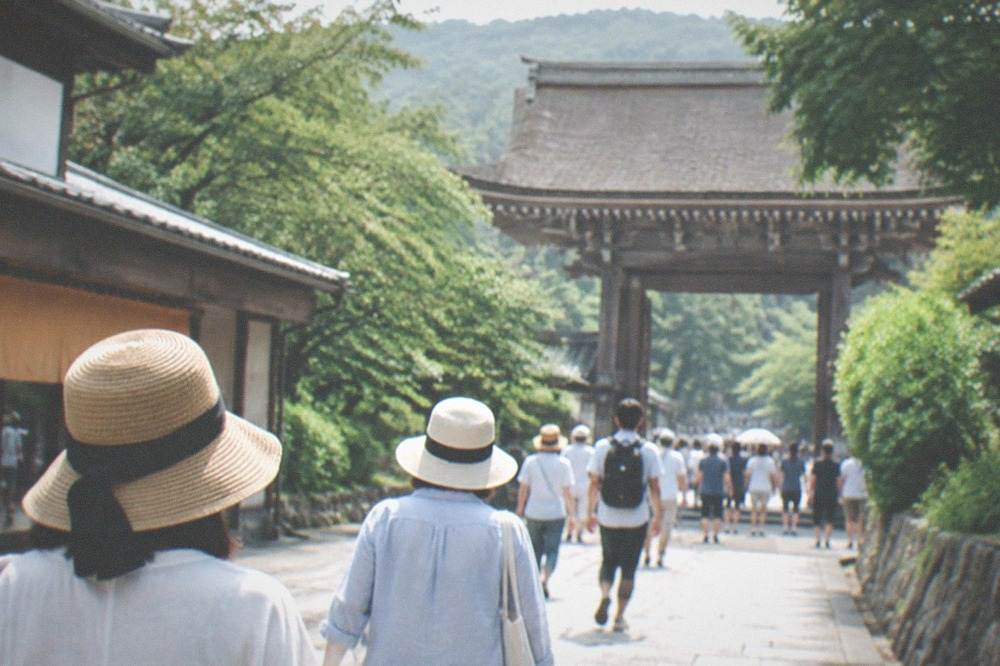 Visitors with sun hats walking a wide stone path toward a large wooden temple gate in bright morning light