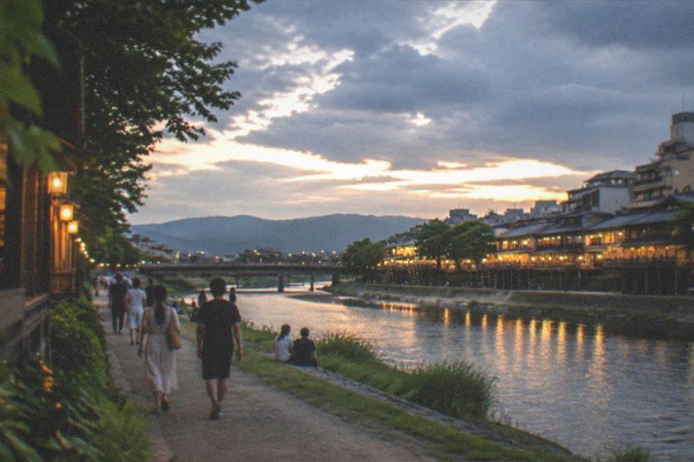 The Kamo River at dusk in summer with people strolling along the bank and restaurants lit on the far side