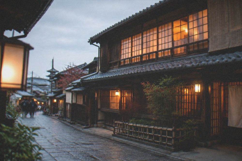 A traditional wooden townhouse at dusk with warm light glowing from its paper-screened windows