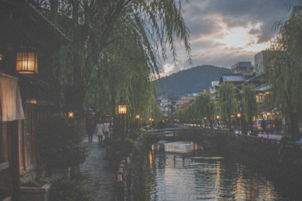 A willow-lined canal street in a traditional onsen town at dusk with lanterns lit