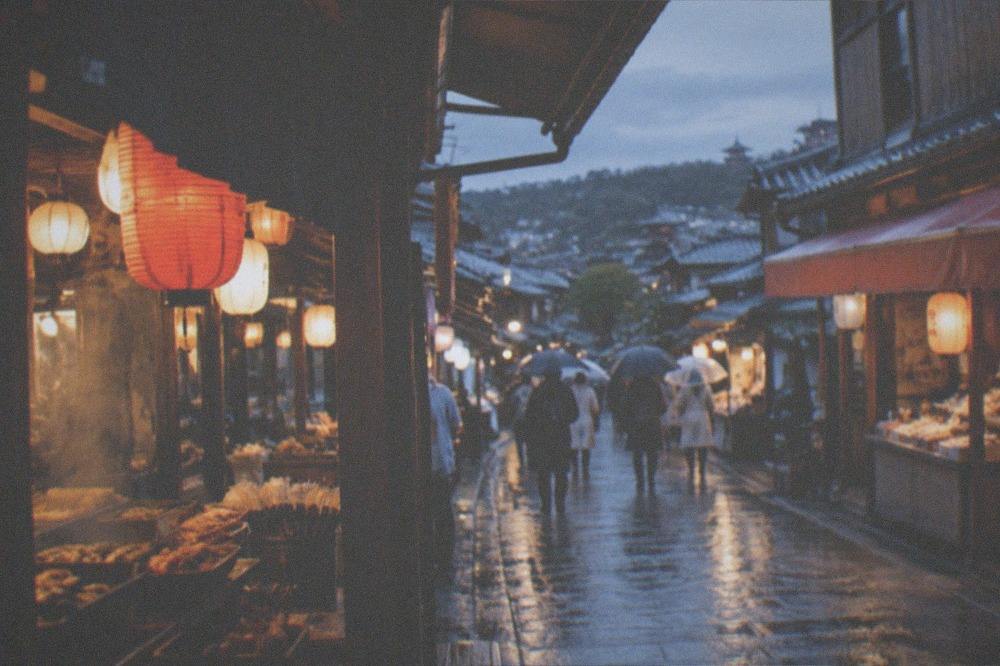 A covered market arcade with food stalls and hanging lanterns glowing against wet evening light