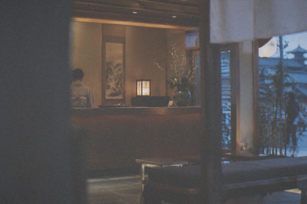 A ryokan reception hall with a wooden counter, hanging scrolls, and a ceramic vase of seasonal flowers