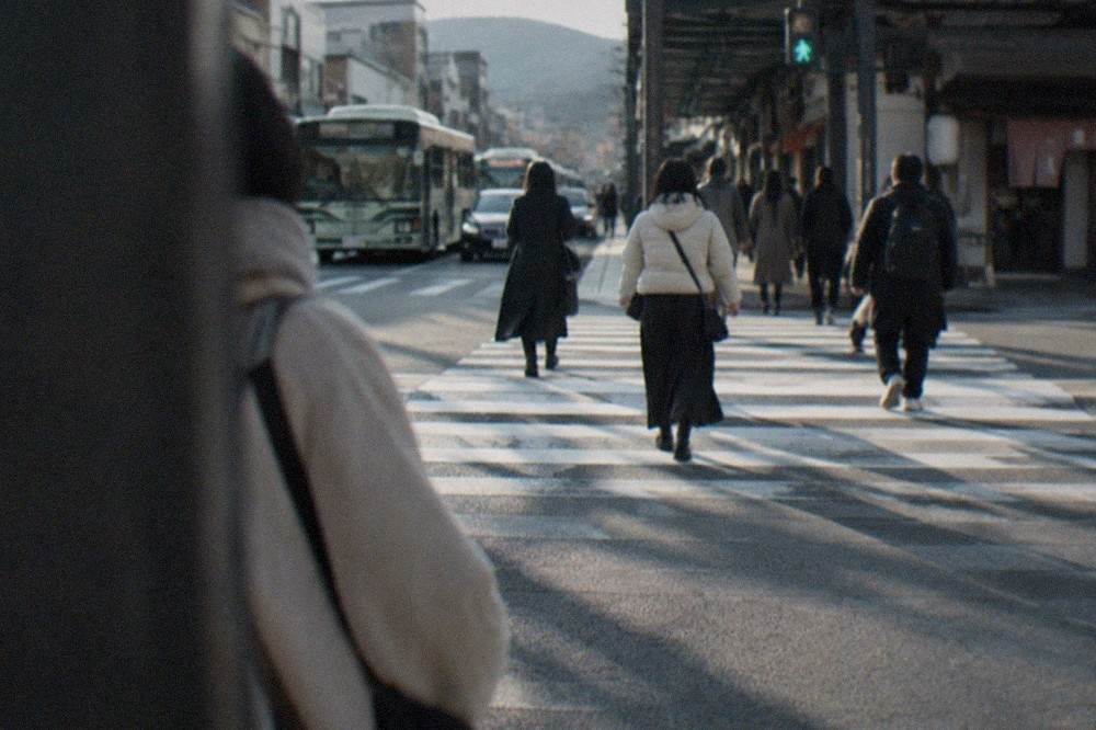 A busy pedestrian crossing on a wide shopping street in Kyoto with buses and shops along both sides