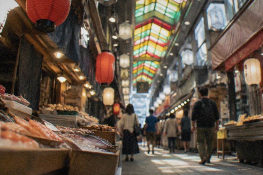A covered market lane lined with small food stalls under lanterns and hanging signs