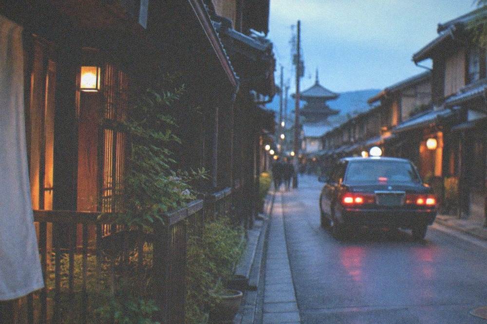 A taxi driving along a narrow street lined with traditional wooden machiya houses in Kyoto