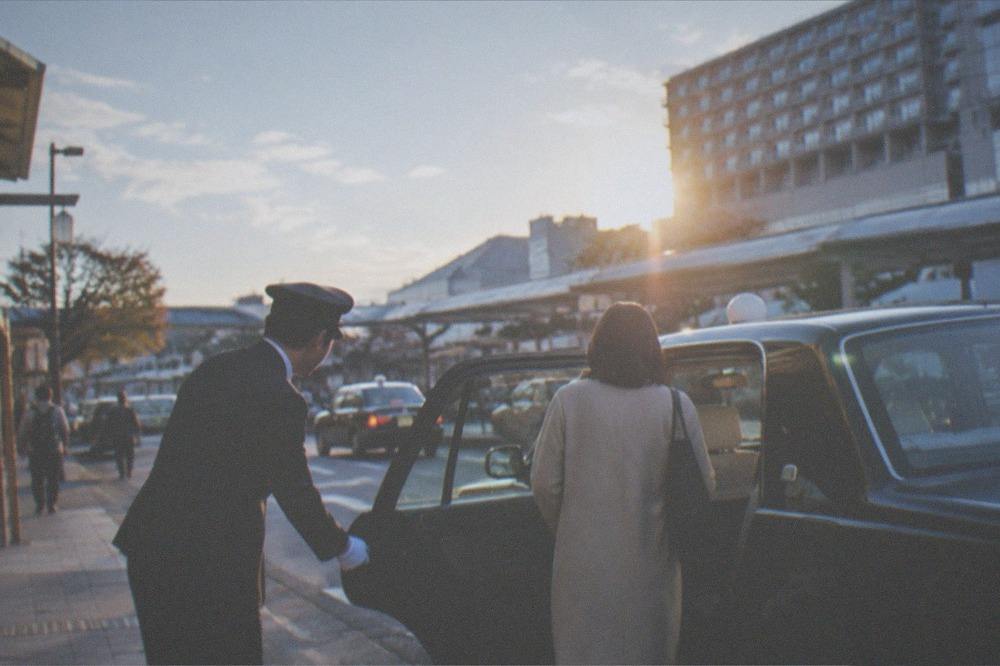 A taxi driver in white gloves and uniform opening the rear passenger door for a customer in Kyoto