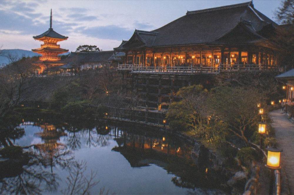 Illuminated temple hall and pagoda reflected in a pond at night with warm lanterns along the walkway