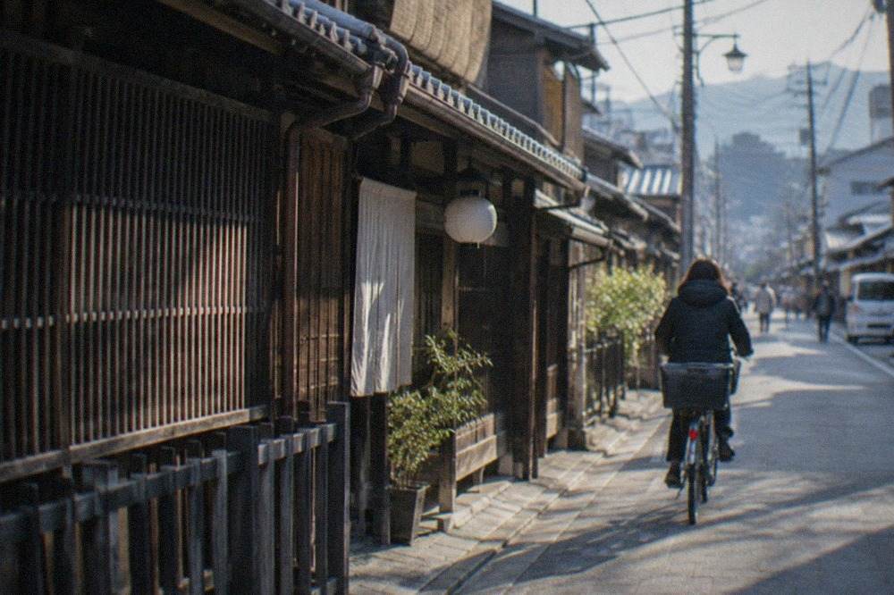 A narrow Kyoto street with traditional wooden machiya facades and a cyclist passing under power lines