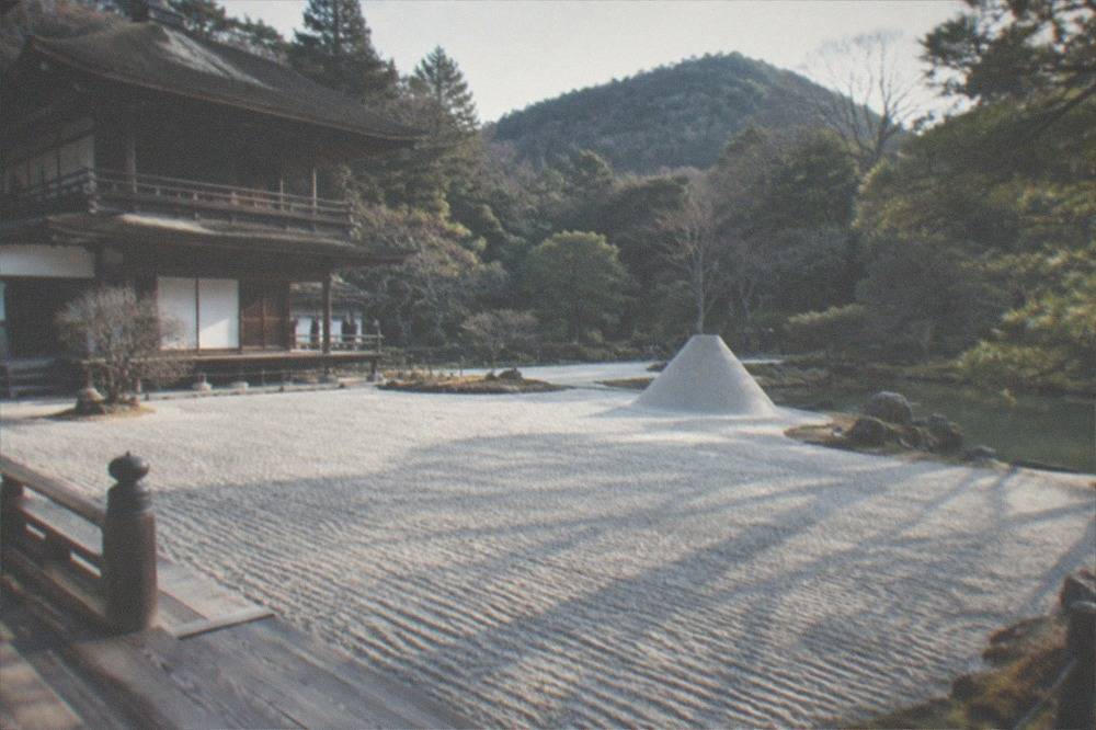 A two-storey wooden pavilion beside a pond, with a wide expanse of raked pale sand and a tall sand cone behind it