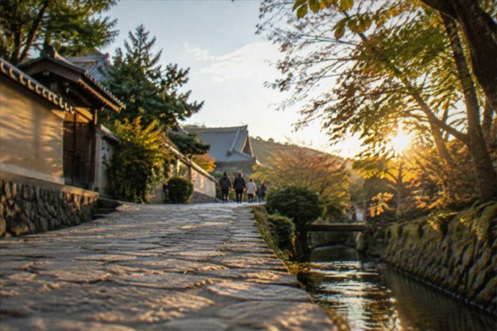 A stone path beside a canal lined with trees leading toward temple rooftops