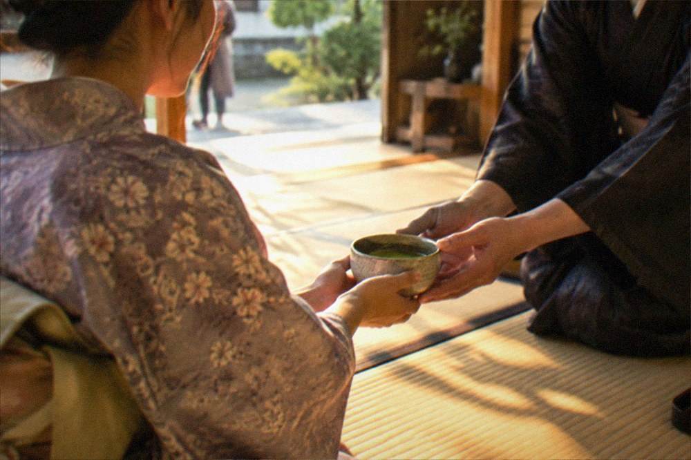 A guest in kimono receiving a bowl of matcha across a tatami floor