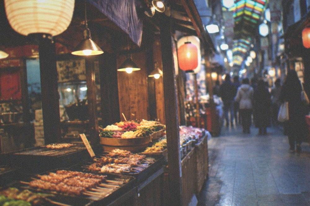 A covered Nishiki Market lane with food stalls selling skewers, pickles and sweets under hanging lanterns