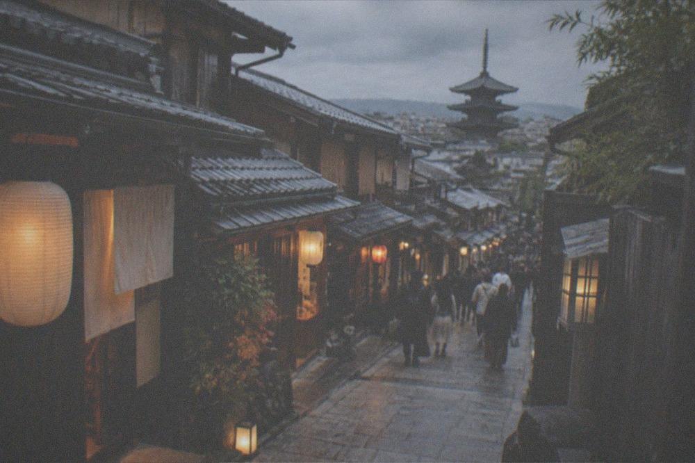 A narrow stone lane between traditional wooden machiya shopfronts lit by lanterns in the early evening