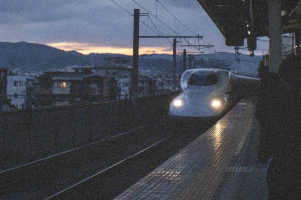 A bullet train platform at dusk with a train's headlights approaching through the gloom