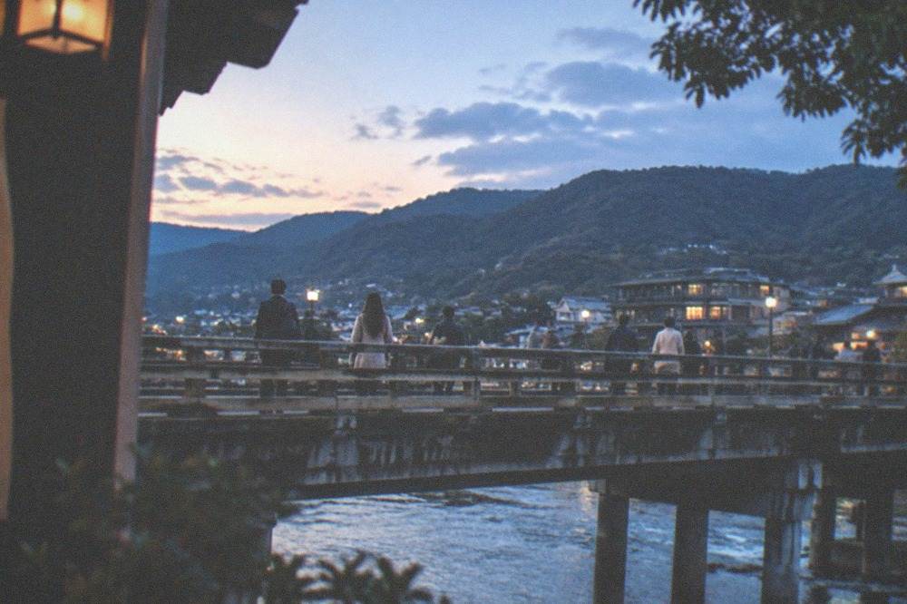 Commuters crossing a bridge over the Kamo River at dusk with the Higashiyama hills behind