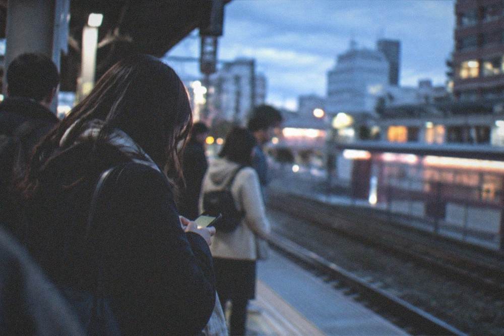 Travelers on a station platform checking a phone beside a departure board showing train times