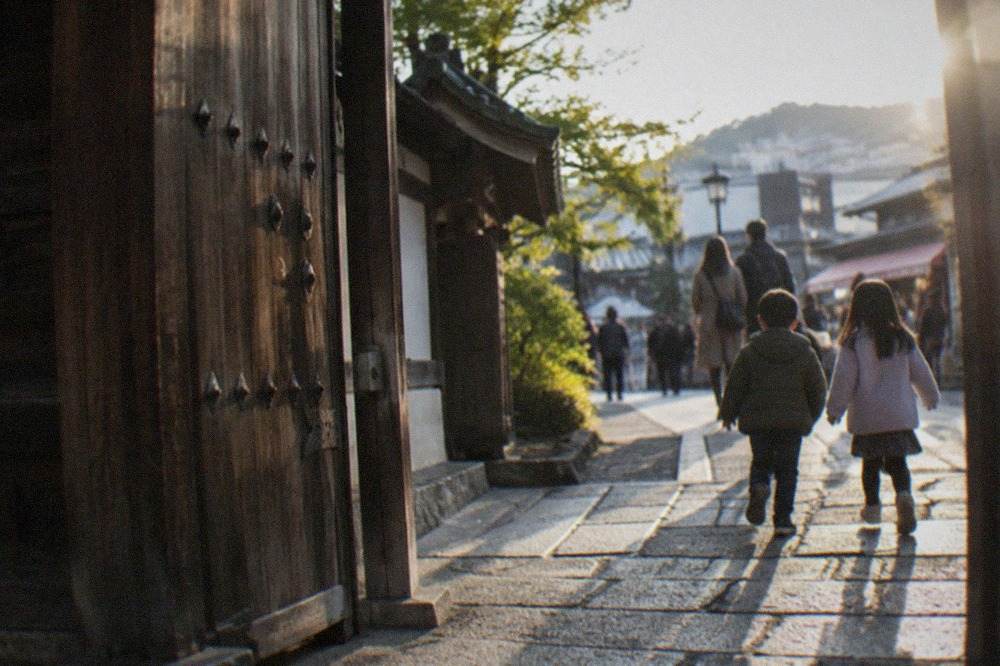 A family walking a stone path beside a wooden temple gate in Kyoto, children leading the way