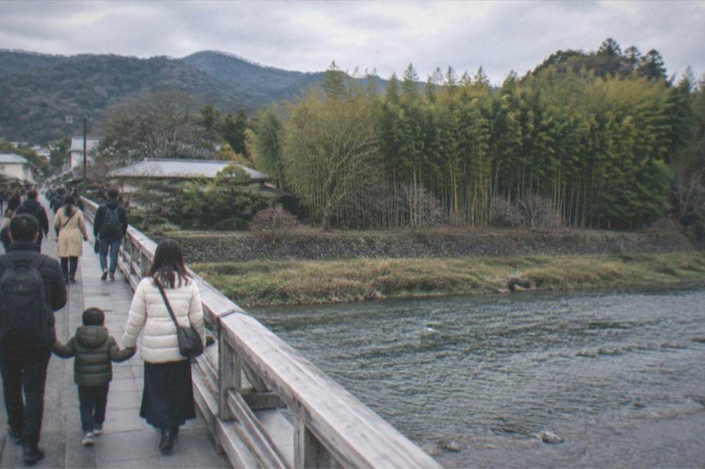 A family crossing a bridge over a river in Arashiyama with bamboo forest on the far bank